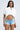 A woman in the SHINE ON BUTTON DOWN BUBBLE TOP, a cropped white bubble-hem shirt, and light blue distressed denim shorts stands against a plain background, confidently facing the camera with long straight hair, glasses, and hoop earrings.