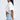 A woman with long straight hair and clear glasses wears the SHINE ON BUTTON DOWN BUBBLE TOP—a loose white cropped top with an elastic bubble hem—and light blue denim shorts, standing in profile against a plain background.