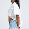 A woman with long straight hair and clear glasses wears the SHINE ON BUTTON DOWN BUBBLE TOP—a loose white cropped top with an elastic bubble hem—and light blue denim shorts, standing in profile against a plain background.