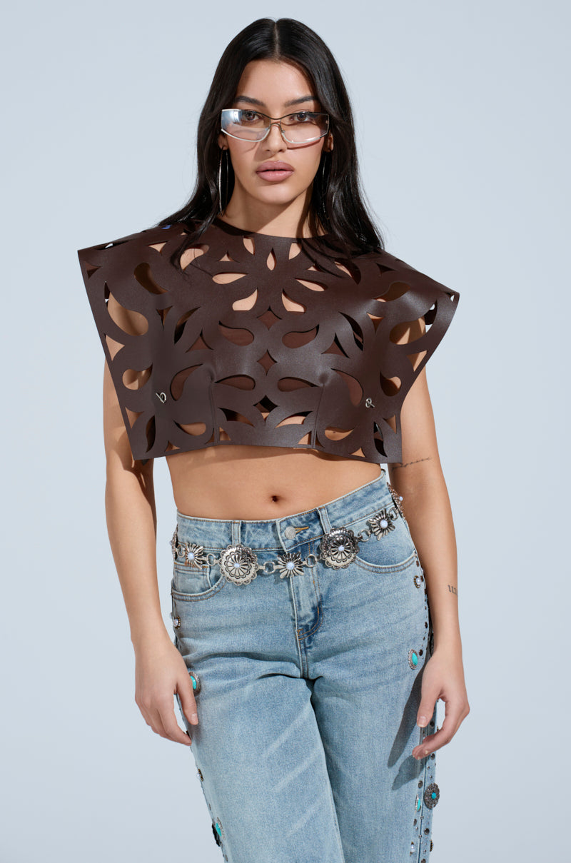 A woman wearing tinted glasses, the LAY LOW CUT OUT BLOUSE in dark brown faux leather, and high-waisted blue jeans with silver conchos stands against a light gray background.