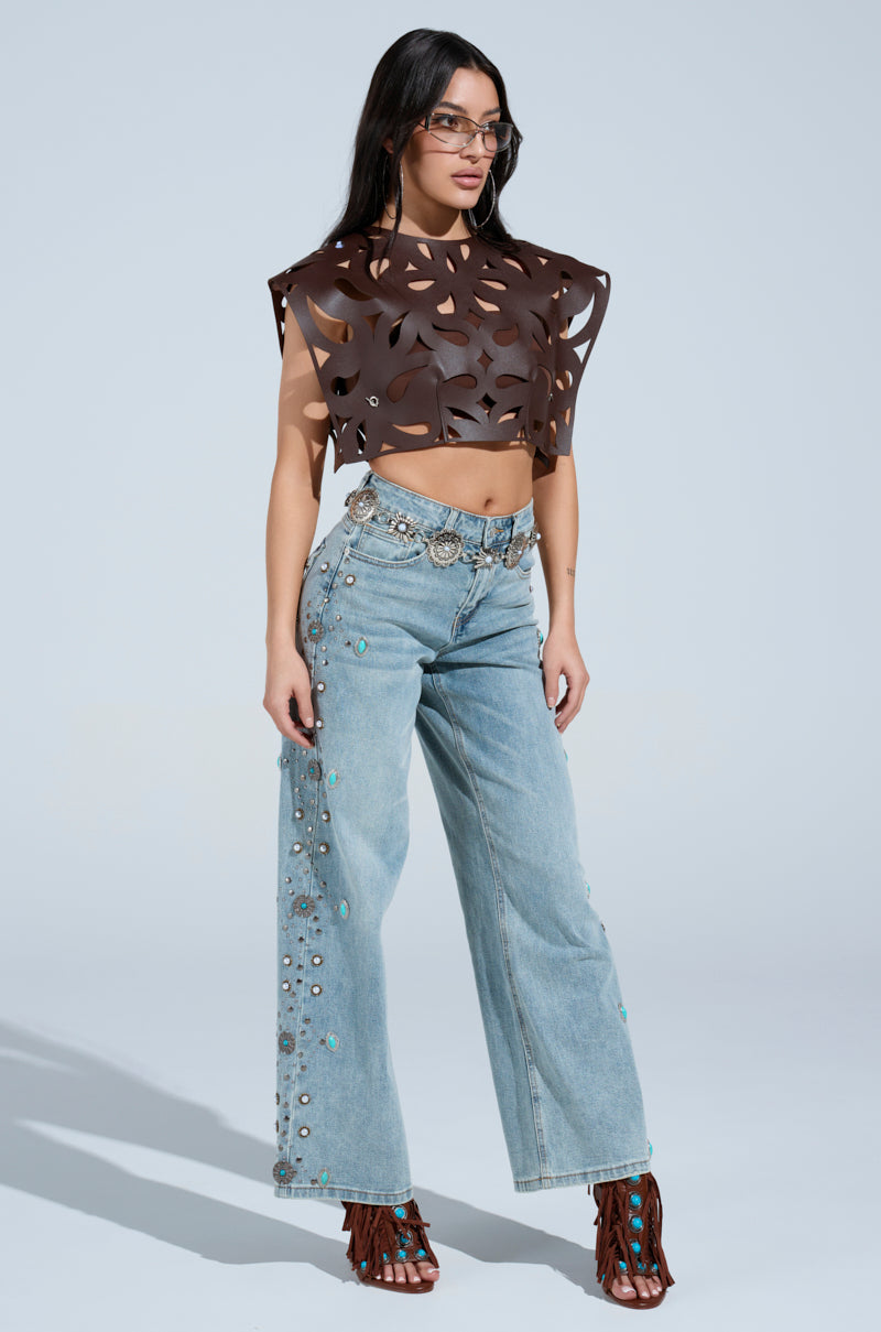 Woman wearing the LAY LOW CUT OUT BLOUSE in brown with geometric cutouts, paired with light blue high-waisted jeans featuring silver grommets and turquoise accents, brown fringed sandals, a silver belt, and sunglasses against a plain background.