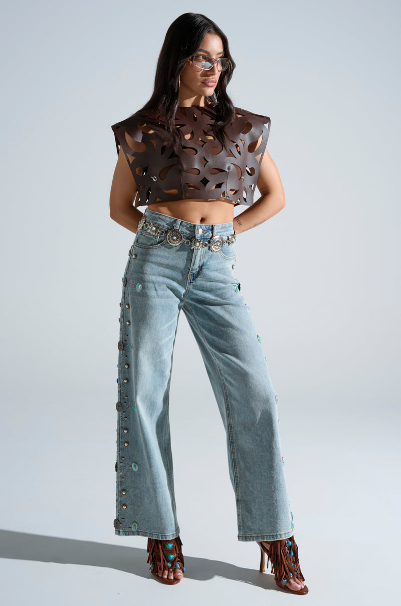 A woman wears the LAY LOW CUT OUT BLOUSE, a brown faux leather crop top with geometric cutouts, paired with light blue high-waisted jeans with side buttons, a silver belt, brown fringed shoes, and sunglasses against a plain background.