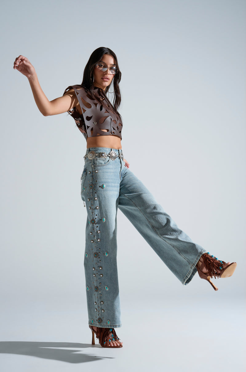 A woman poses confidently in the LAY LOW CUT OUT BLOUSE, styled with embellished light blue jeans with side grommets and brown lace-up heels. She accessorizes with sunglasses and jewelry, standing on one leg against a plain gray background.