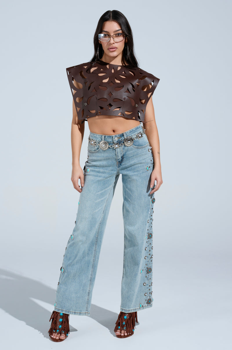A woman poses against a light background wearing the LAY LOW CUT OUT BLOUSE, a brown crop top with geometric cutouts, paired with light blue high-waisted embellished jeans, a silver belt, brown fringe sandals, and glasses.