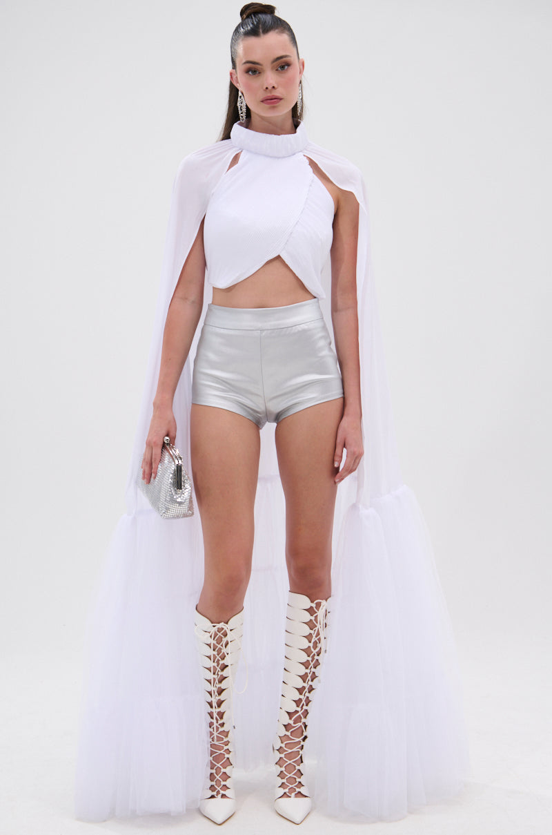 A woman wears the DONT WASTE MY TIME BLOUSE—a white mock neck crop top with voluminous gathered sleeves and a sheer cape—paired with silver high-waisted shorts, tall white lace-up boots, and a small silver handbag against a white background. 1