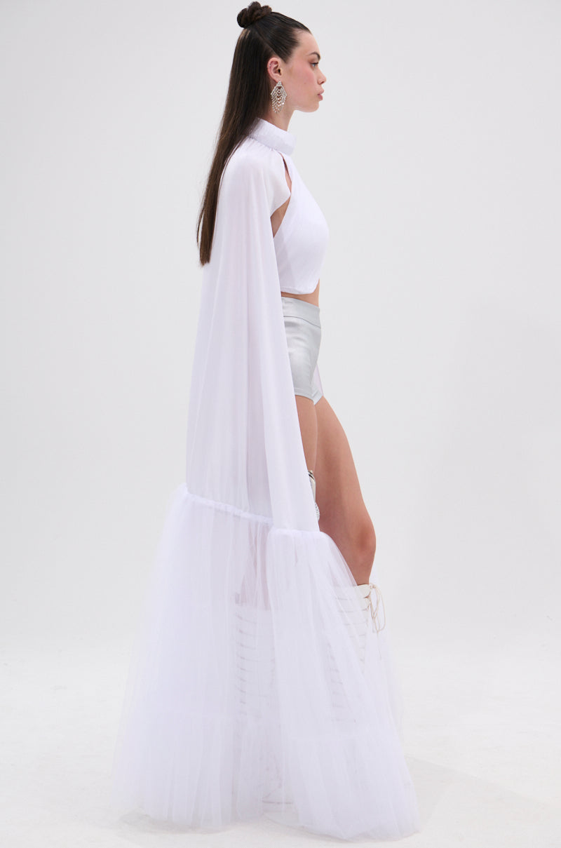 A woman in profile wears the DONT WASTE MY TIME BLOUSE, a white sleeveless mock neck crop top, paired with high-waisted silver shorts, a sheer white tulle cape with ruffled hem, hoop earrings, lace-up knee-high boots, and long straight hair. 5