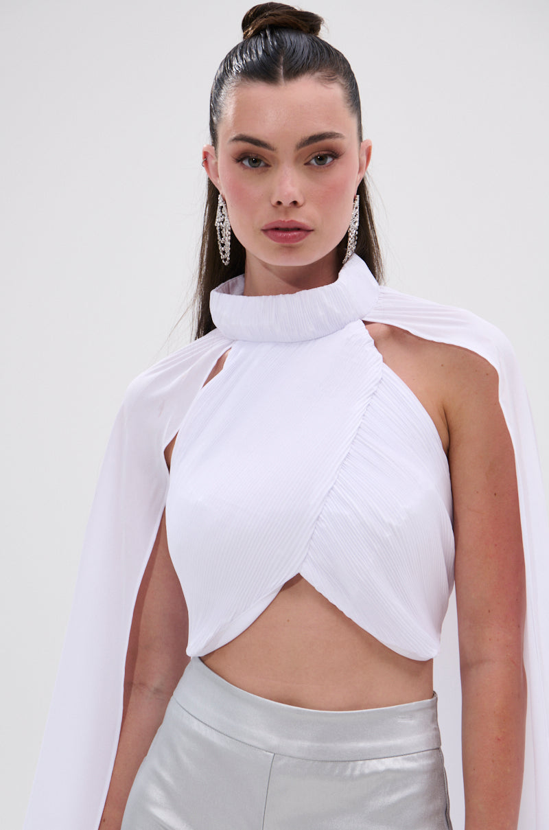 A woman with long dark hair in a high bun wears the sleeveless, mock neck DONT WASTE MY TIME BLOUSE in white with a cape detail, metallic silver high-waisted pants, and long earrings against a plain background. 7
