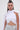A woman with long dark hair in a high bun wears the sleeveless, mock neck DONT WASTE MY TIME BLOUSE in white with a cape detail, metallic silver high-waisted pants, and long earrings against a plain background.