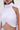 A woman with long brown hair and dangling earrings wears the DONT WASTE MY TIME BLOUSE—a sleeveless, sheer white mesh top featuring a high neck, pleated detail, draped asymmetry, and midriff-baring design.