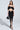 A woman with long straight hair and a neutral expression models the DREAM CATCHER MAXI SKIRT IN BLACK in a studio, paired with strappy heels and a black handbag, showing off its curve-hugging fit, deep V cutout, and high slit.