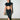 A woman with long dark hair in a high ponytail wears a black crop top and the DREAM CATCHER MAXI SKIRT IN BLACK, featuring a sultry front slit, holding a small clear purse against a white brick wall on polished concrete.
