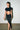 A woman with long dark hair in a high ponytail wears a black crop top and the DREAM CATCHER MAXI SKIRT IN BLACK, featuring a sultry front slit, holding a small clear purse against a white brick wall on polished concrete.