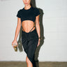 A woman with long dark hair in a high ponytail wears a black crop top and the DREAM CATCHER MAXI SKIRT IN BLACK, featuring a sultry front slit, holding a small clear purse against a white brick wall on polished concrete.