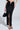 A person wears the DREAM CATCHER MAXI SKIRT IN BLACK with a front slit, paired with black strappy heels and a black handbag with a gold handle. The image is cropped to show only the lower torso and legs.
