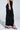 A woman stands sideways against a plain light background, wearing the curve-hugging DREAM CATCHER MAXI SKIRT IN BLACK paired with black high-heeled sandals.