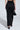 A woman stands facing away in the DREAM CATCHER MAXI SKIRT IN BLACK with a curve-hugging fit, styled with black high-heeled sandals and a black handbag with gold handles, set against a plain light background.