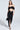 A woman poses against a plain backdrop wearing the DREAM CATCHER MAXI SKIRT IN BLACK, paired with a matching off-the-shoulder top. She accessorizes with a small black handbag and strappy black heels for a chic, curve-hugging look.
