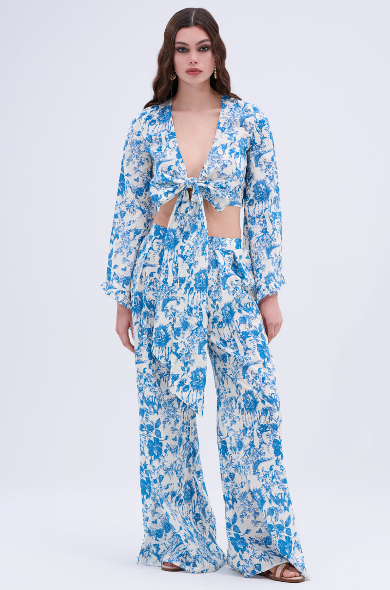 A woman with long, wavy brown hair and a neutral expression stands against a plain background wearing the DOLLED UP PALAZZO PANT—a vintage floral print set featuring a deep V-neck, tie-front top with cutouts, and high-waisted palazzo pants.