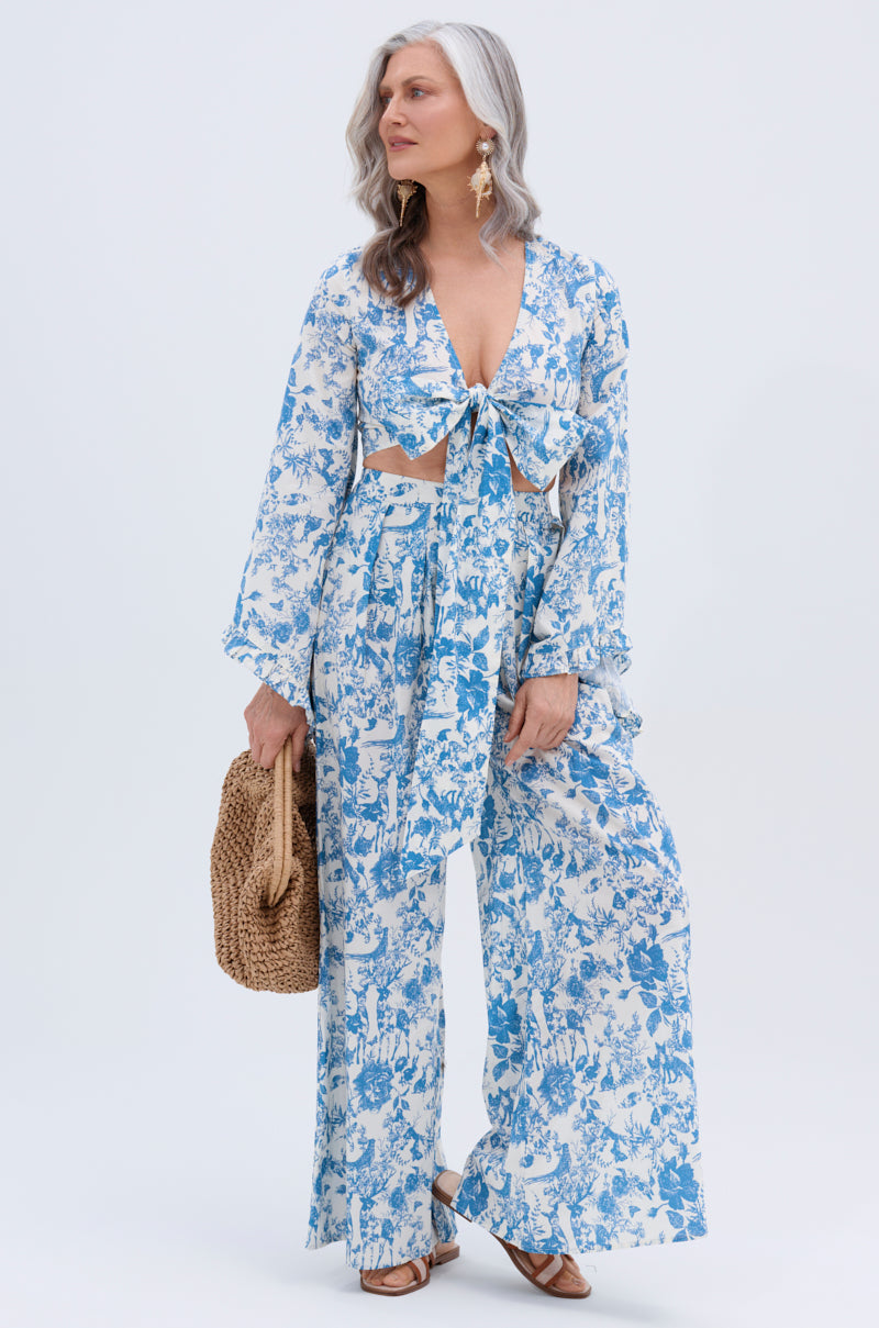 A woman with long gray hair models the DOLLED UP PALAZZO PANT, a blue and white vintage floral two-piece outfit, paired with a woven bag and brown sandals against a plain light background.