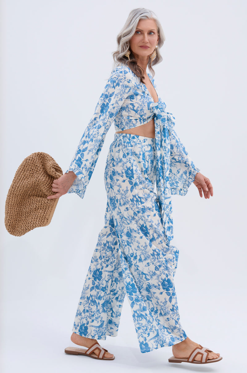 A woman with long gray hair wears the DOLLED UP PALAZZO PANT in a blue and white vintage floral print, paired with brown sandals and holding a woven straw hat, walking against a plain light background.