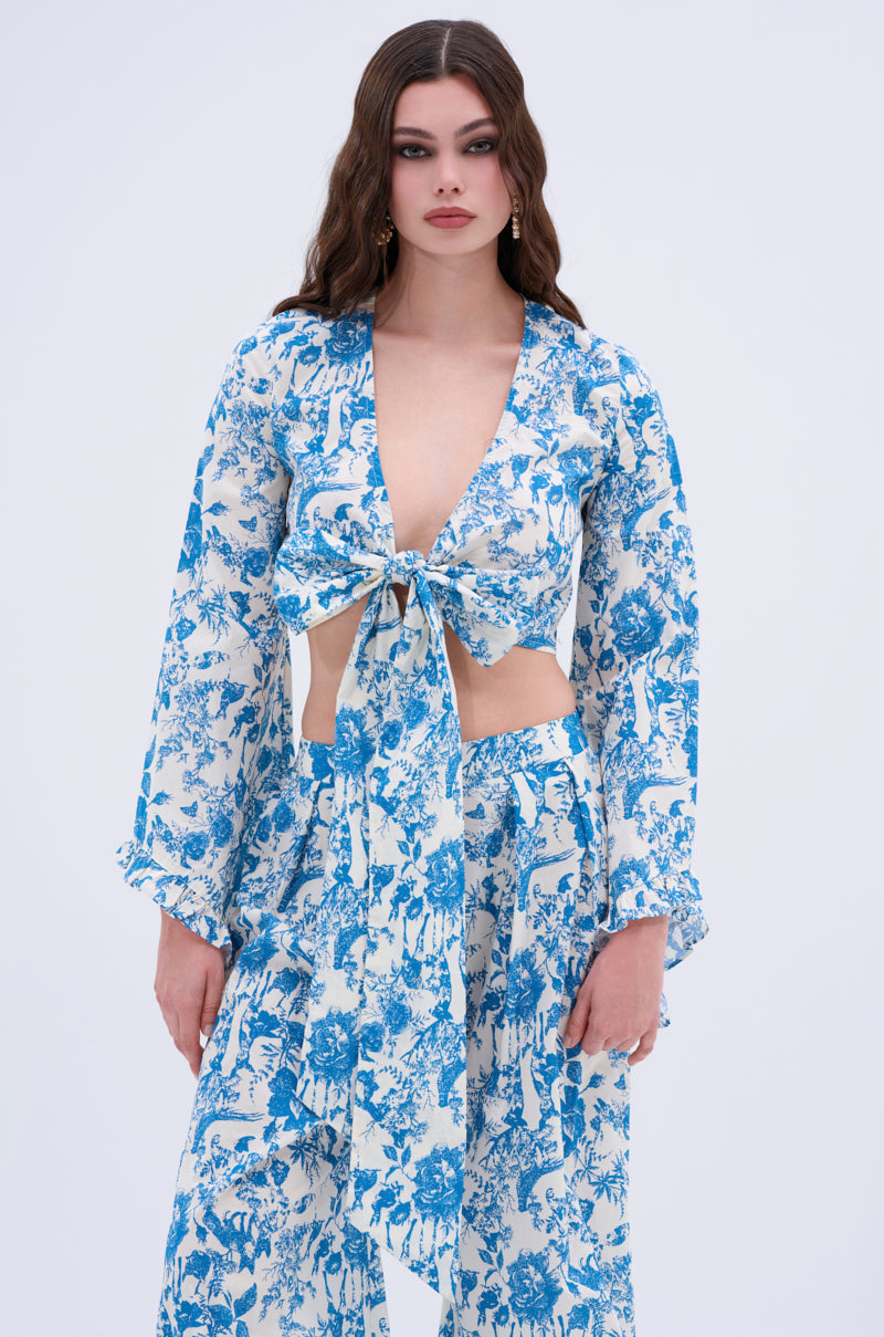 A woman with long wavy brown hair wears the DOLLED UP TIE FRONT TOP in blue and white vintage floral print, paired with matching wide-leg pants, standing against a plain light background.