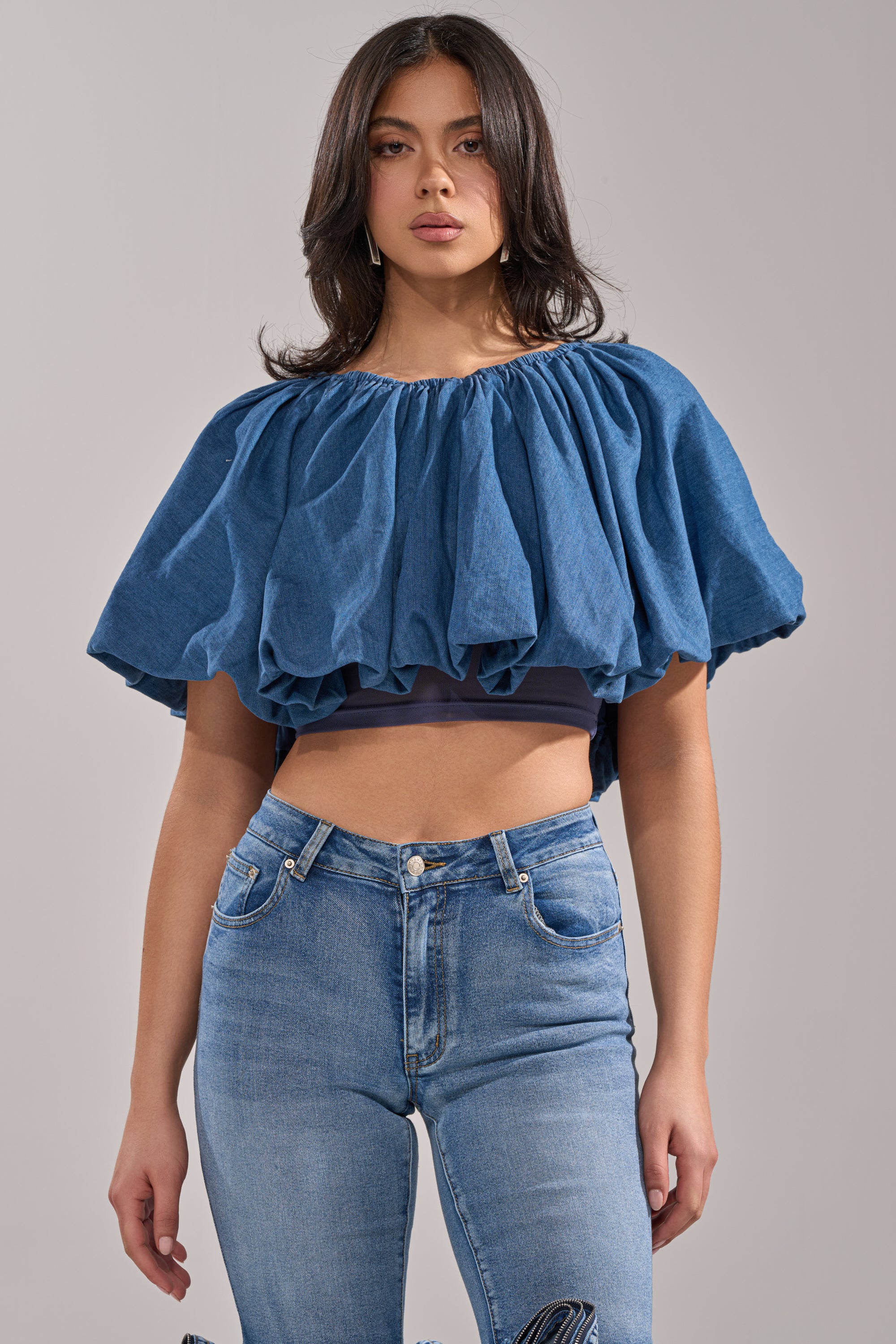 A woman wears the NEVER GOING TO STOP BLOUSE IN MEDIUM BLUE DENIM, an oversized cropped blouse with puffed sleeves, paired with high-waisted blue jeans, standing against a plain light background.