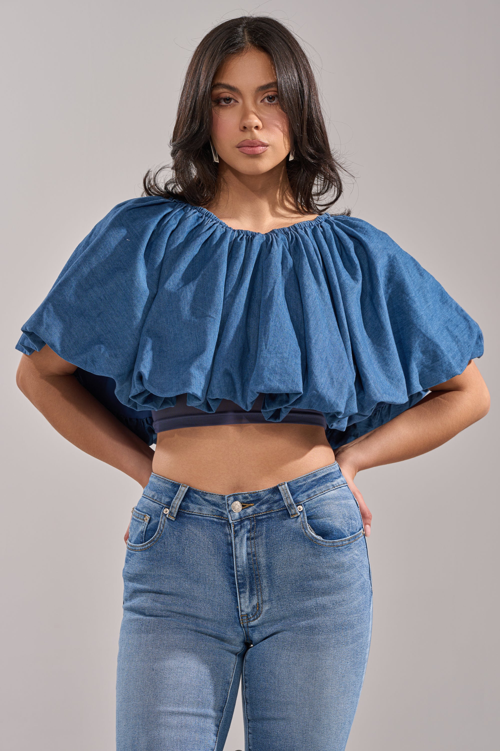 A woman models the NEVER GOING TO STOP BLOUSE IN MEDIUM BLUE DENIM, a voluminous, ruffled cropped top, paired with high-waisted blue jeans and poses confidently against a neutral background to showcase its trendy oversized look.