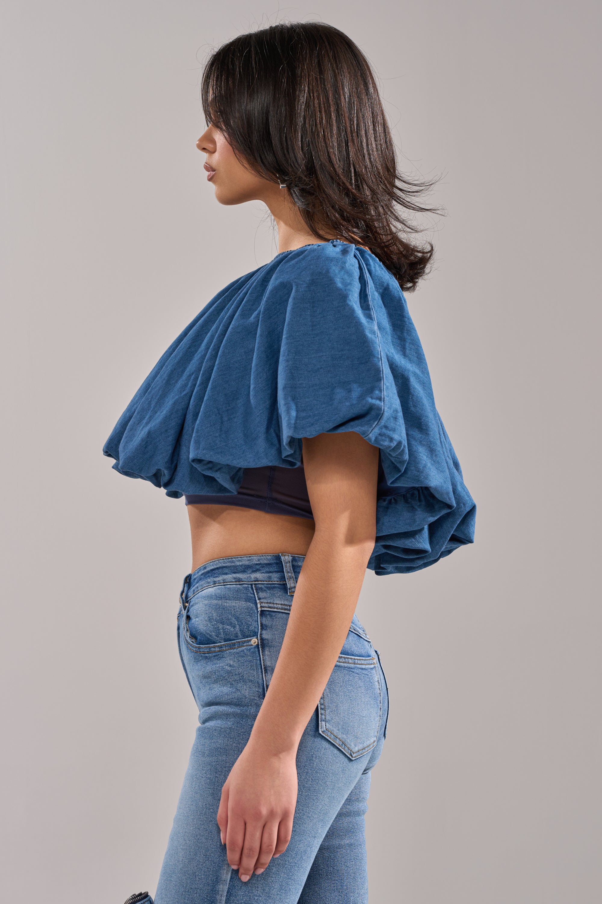A woman stands in profile wearing the NEVER GOING TO STOP BLOUSE IN MEDIUM BLUE DENIM, featuring a loose, oversized fit and short puffed sleeves, paired with fitted blue jeans against a plain light gray background.