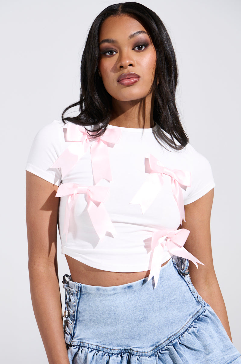 A person with long black hair wears the PRETTY PLEASE TEE, a white cropped top with satin bows, paired with a light blue ruched denim skirt. They pose against a plain, light-colored background.