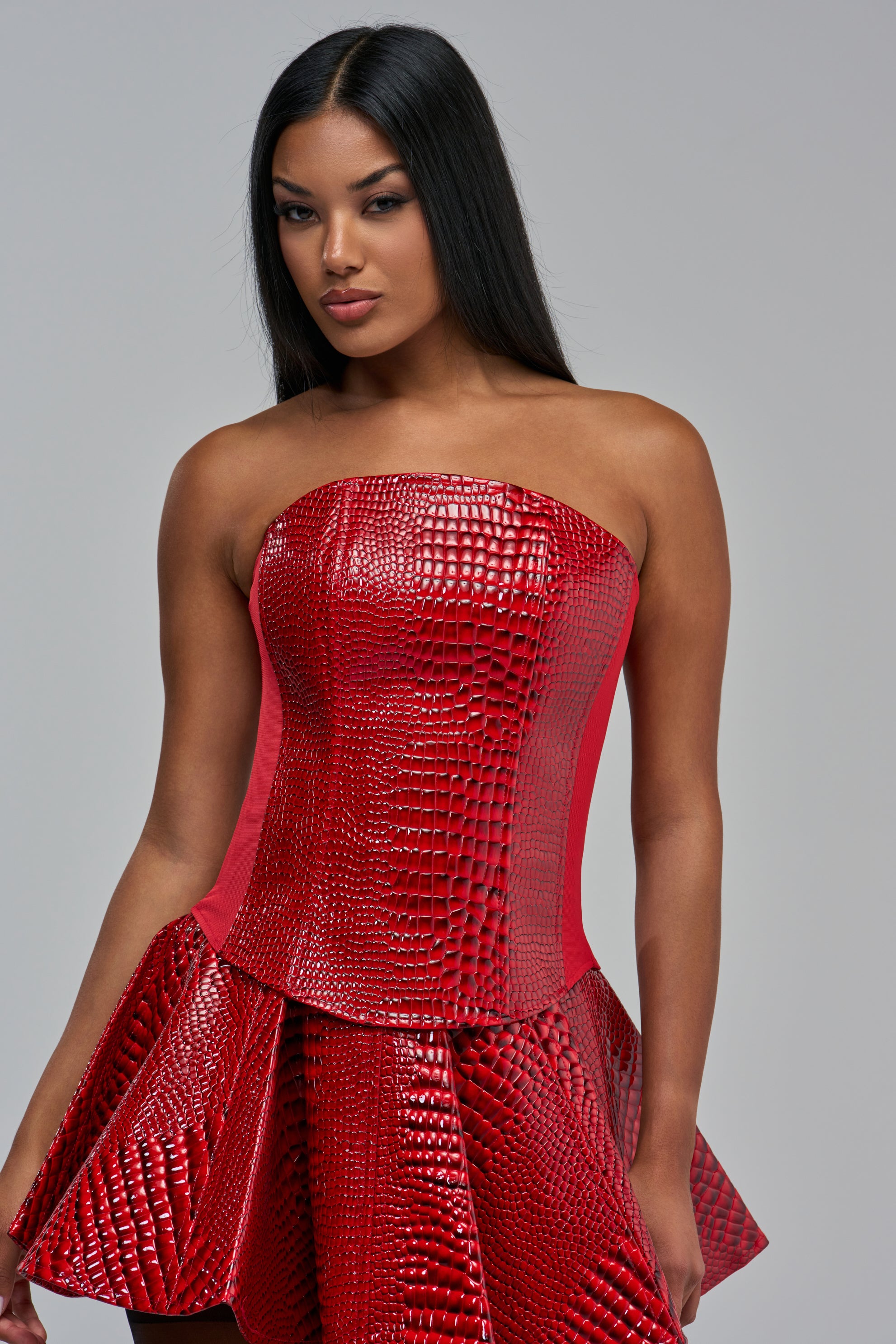 A model wears the CAROLINE CROC TOP, a red faux patent leather corset with an embossed crocodile pattern, paired with a matching flared skirt.