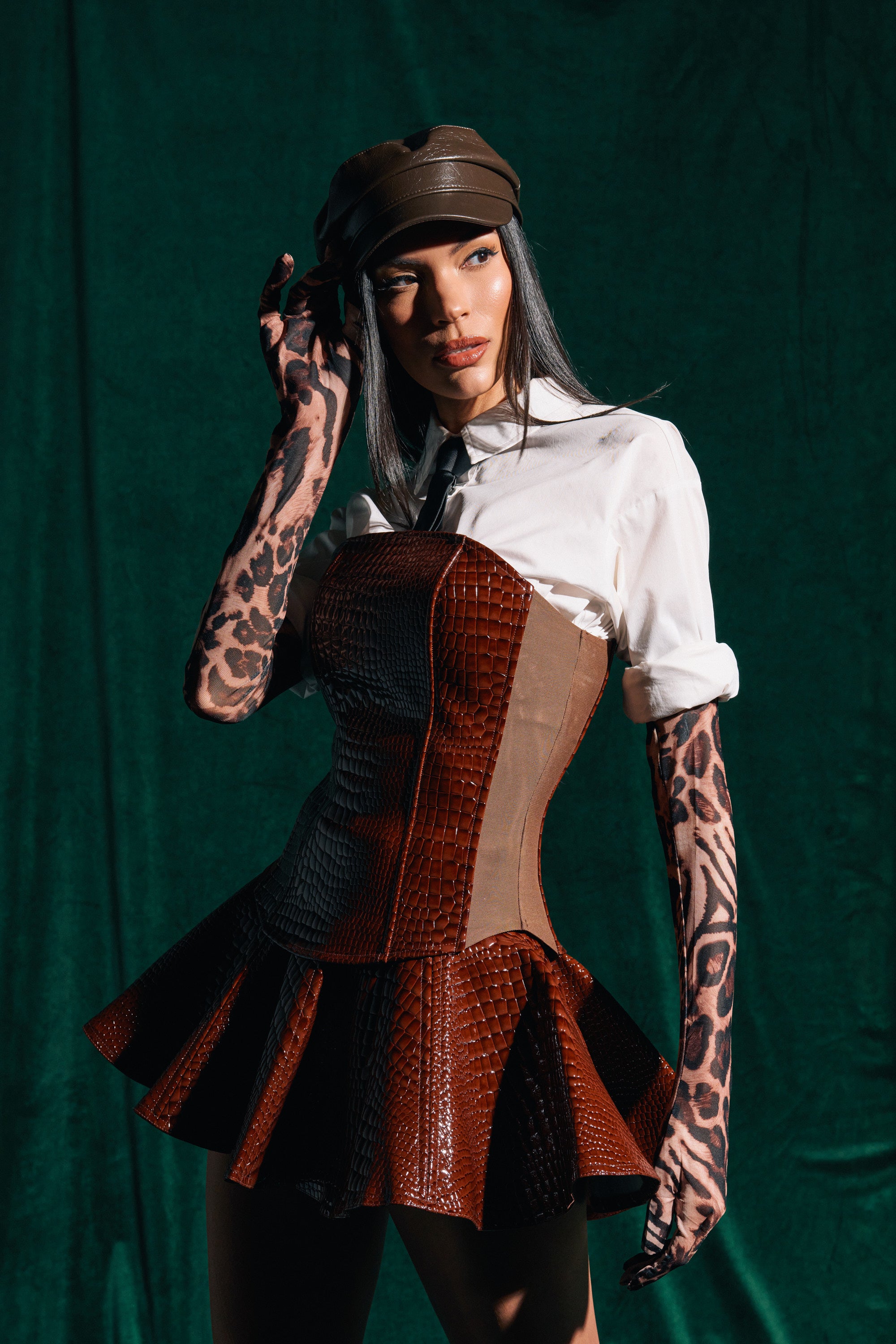 A woman poses against a dark green background wearing the TOO GOOD FOR YOU CROCODILE CORSET over a white collared shirt, styled with a brown leather hat and animal print gloves as she touches her hat and gazes to the side.