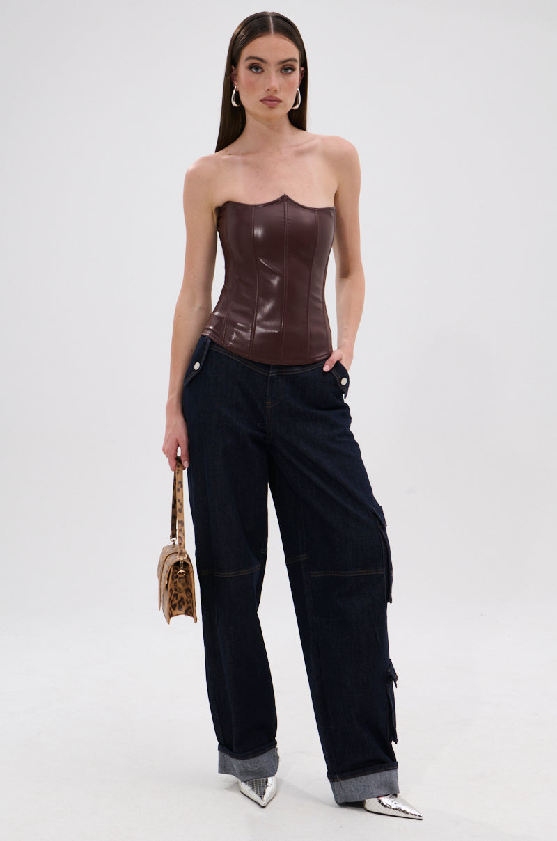 A woman poses against a plain background in the WILD WEST CORSET TOP, featuring a strapless faux leather design with a sweetheart neckline, styled with cuffed dark wide-leg jeans, silver pointed shoes, hoop earrings, and an animal print handbag. 5