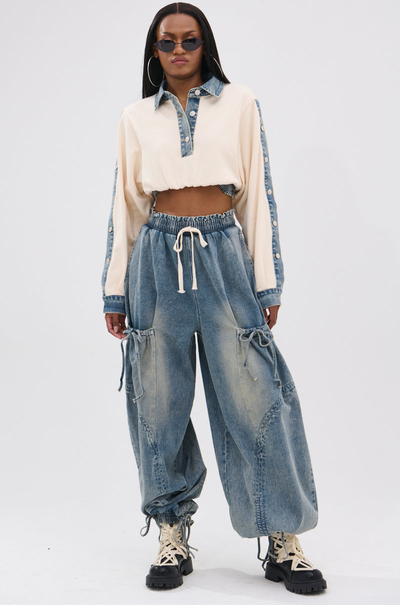 A woman poses in RELAX WITH ME JOGGER vintage-washed denim jeans with drawstrings and side pockets, a cropped cream and denim button-up top, chunky beige boots, dark sunglasses, and hoop earrings against a plain background.
