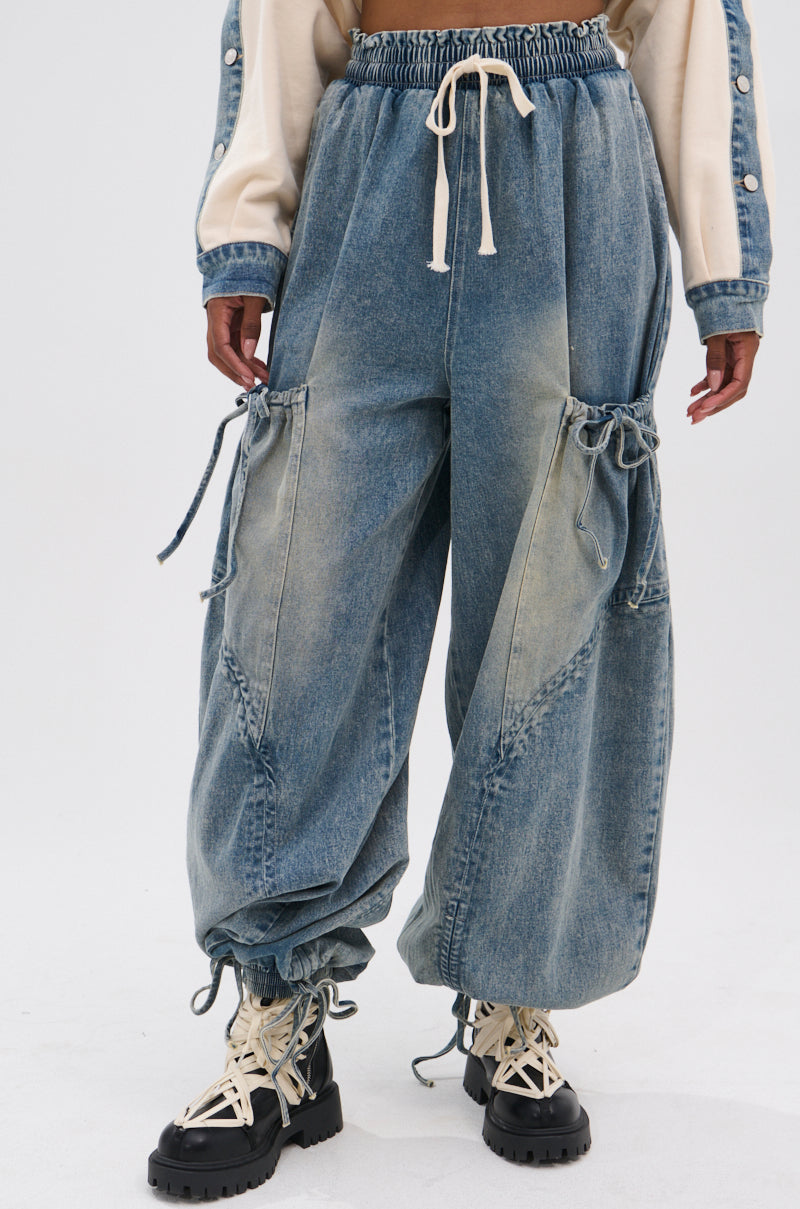 A person wears the RELAX WITH ME JOGGER, a vintage-washed, high-rise denim cargo jogger with drawstrings at the waist and ankles, styled with chunky lace-up boots and a beige long-sleeve top featuring button details.