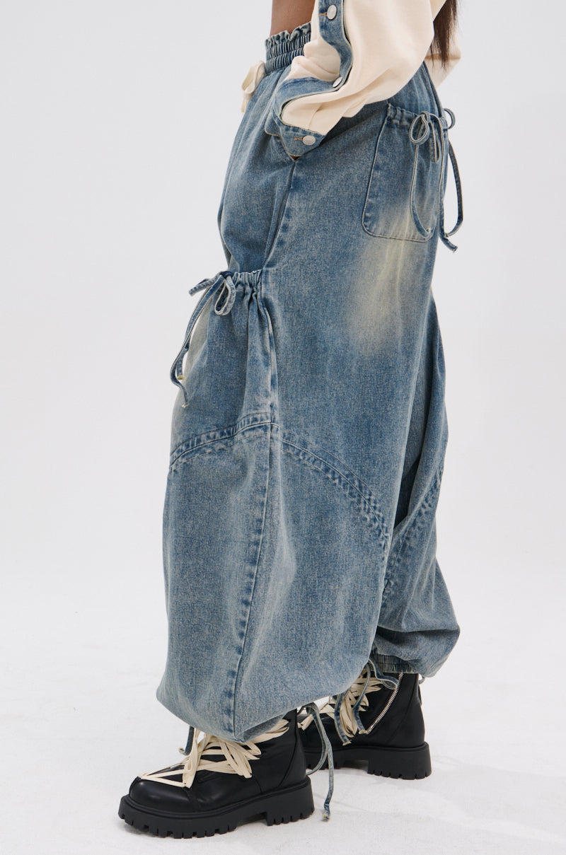 Wearing the RELAX WITH ME JOGGER, featuring loose, vintage-washed denim blue fabric, a high-rise fit, and drawstring details, paired with black lace-up boots, someone poses against a plain light background.