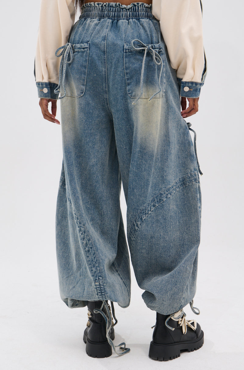 A person is seen from behind wearing the RELAX WITH ME JOGGER in vintage-washed denim with back pockets and drawstring details, paired with black chunky boots and a cream-colored long-sleeve top.