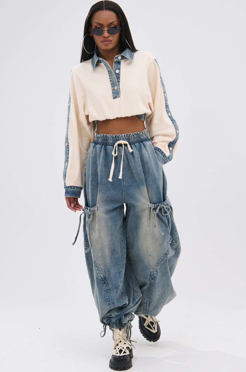 A woman in sunglasses, a cropped cream and denim shirt, and RELAX WITH ME JOGGER pants with a high-rise fit and large pockets strides confidently towards the camera against a plain background.
