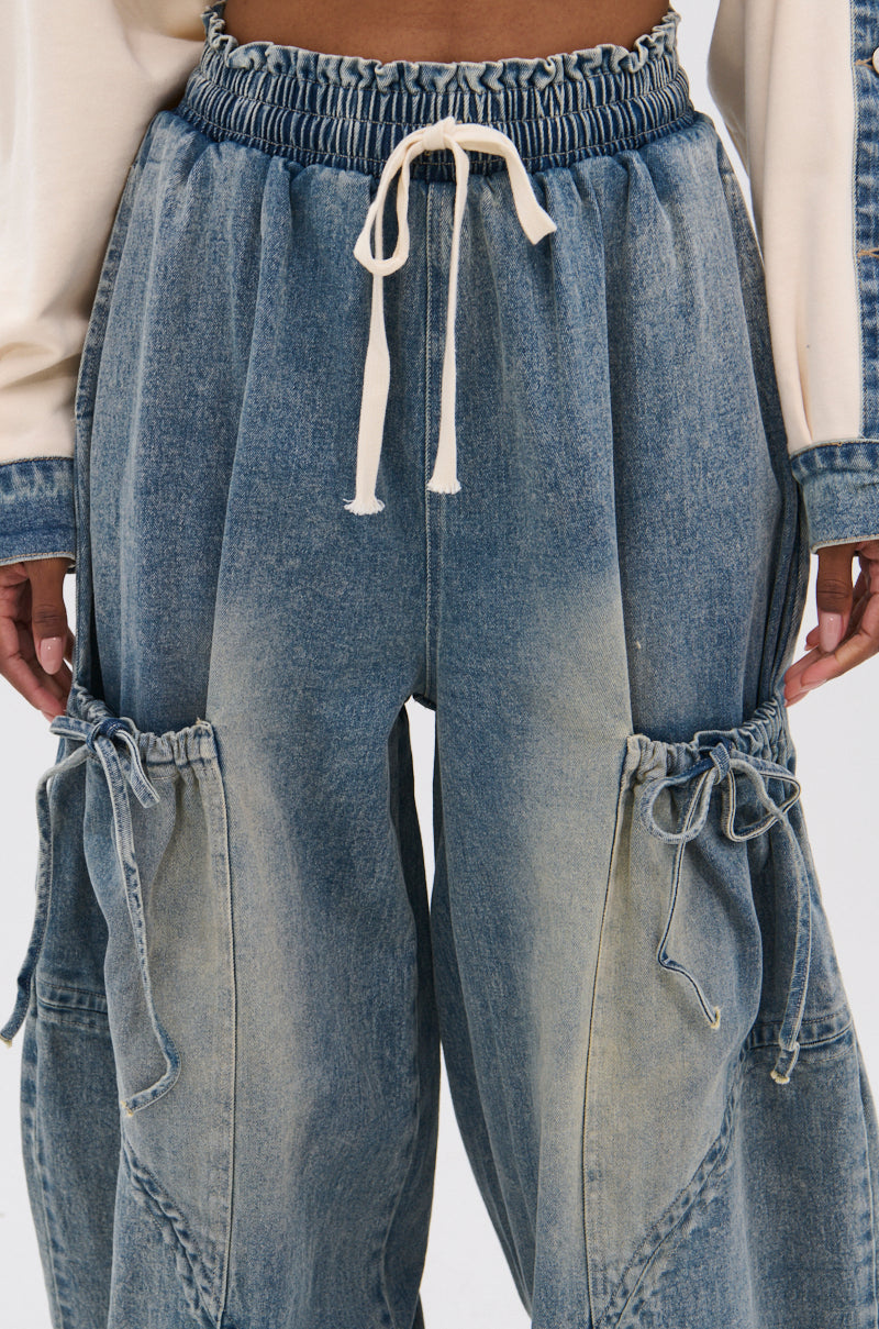 A close-up of a person wearing the RELAX WITH WITH ME JOGGER—vintage-washed denim cargo pants with drawstring waist, side pockets, and a high-rise fit—perfectly paired with a long-sleeve top. Hands are partially visible.
