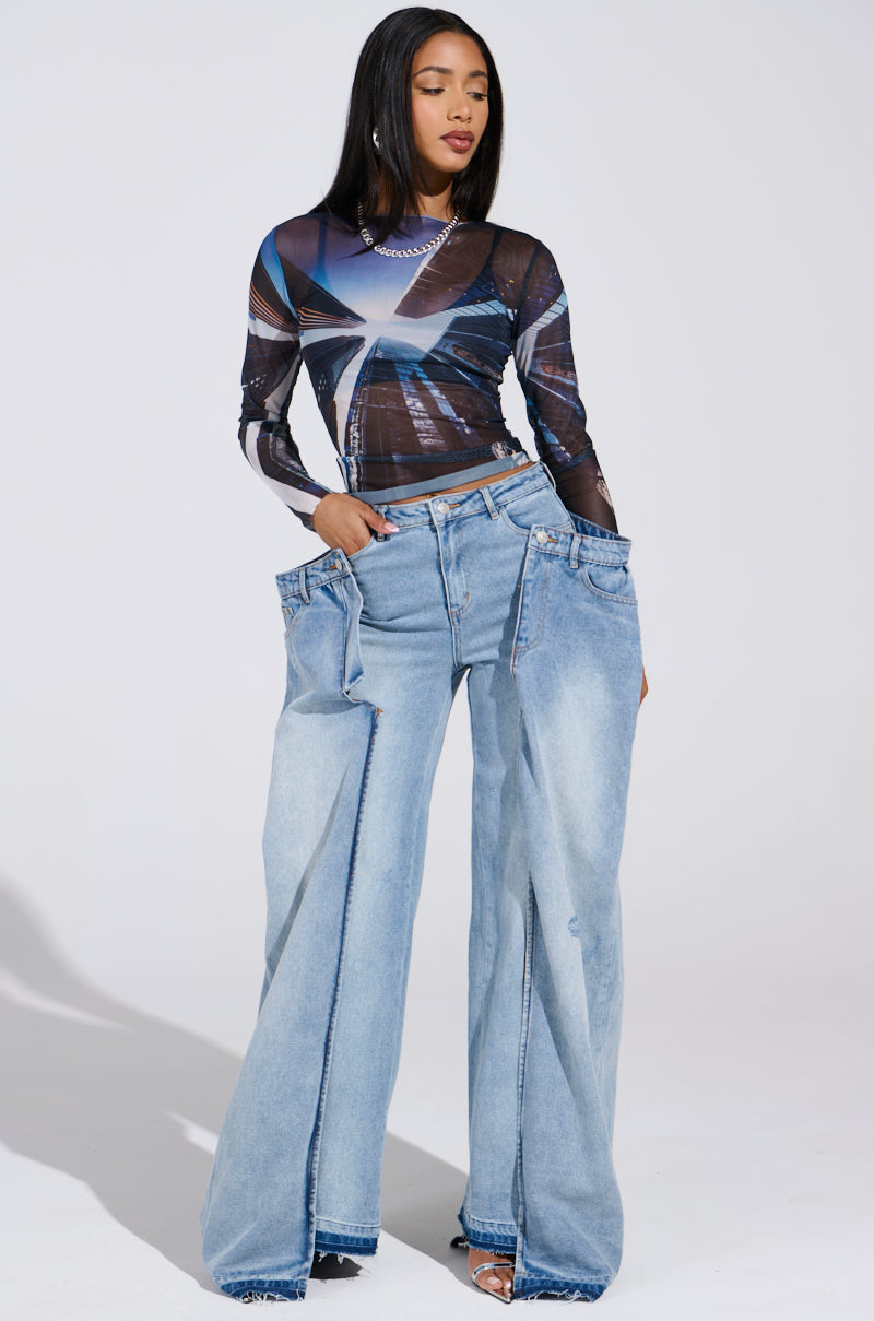 A woman confidently poses in a sheer geometric top and SEEING DOUBLE DECONSTRUCTED DENIM JEANS with oversized light blue high-rise fit. Against a plain white background, her sleek long hair and subtle jewelry complete her fashionable look.