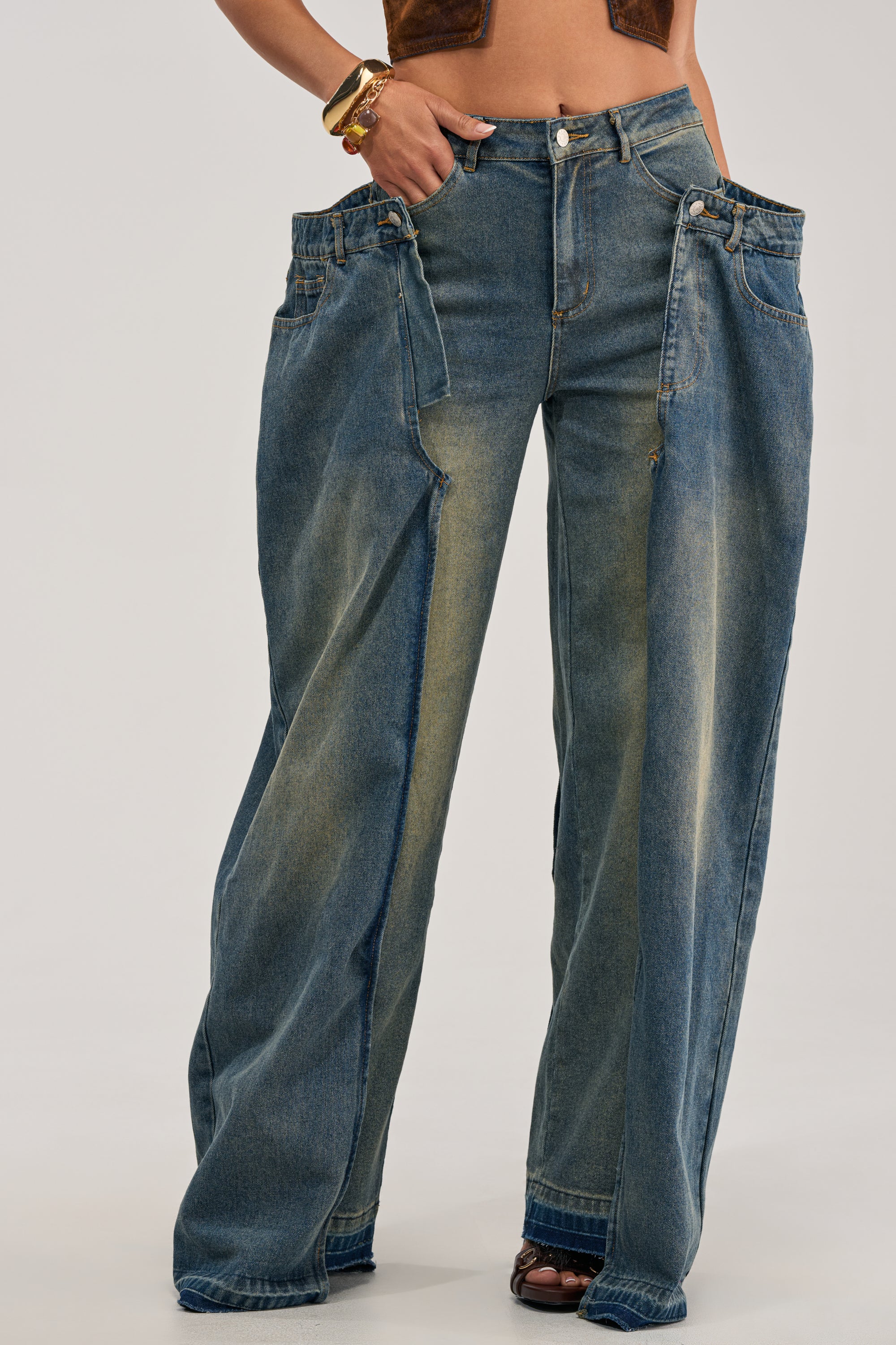 A person wearing the SEEING DOUBLE DECONSTRUCTED VINTAGE JEANS, featuring streetwear-inspired wide-leg denim with double waistbands and belt loops, paired with a brown crop top. Only the lower torso and legs are visible against a plain background.