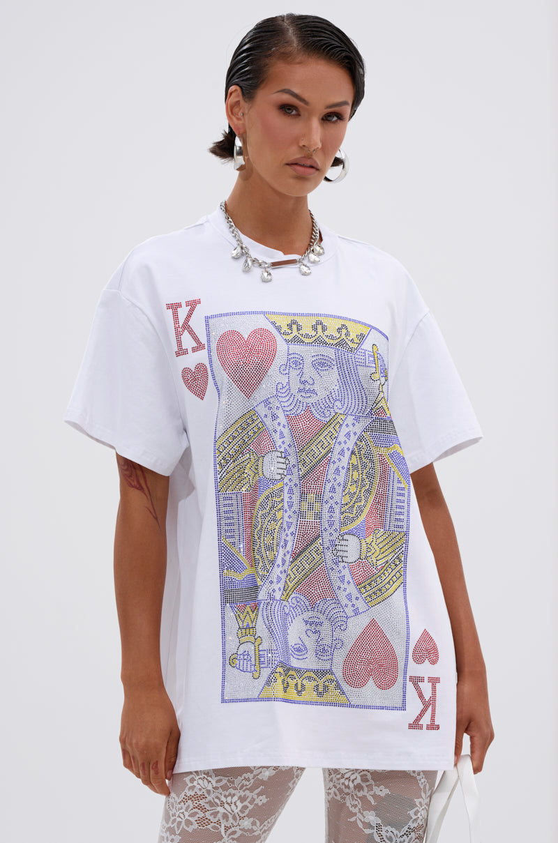 A woman with slicked-back hair wears the KING OF HEARTS OVERSIZED TEE IN WHITE, which features a bold playing card graphic, styled with silver jewelry and white lace leggings.