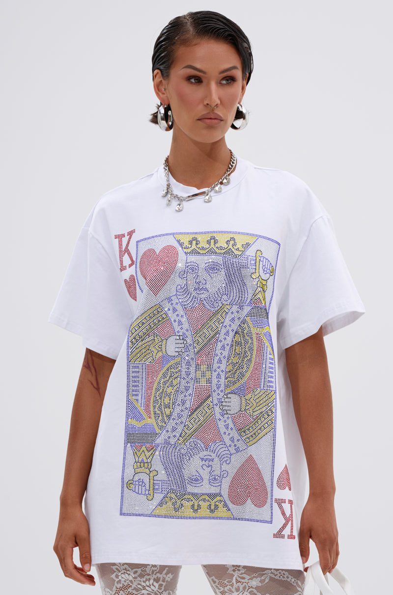 Woman wearing the KING OF HEARTS OVERSIZED TEE IN WHITE, a rhinestone graphic t-shirt, styled with lace pants, silver hoop earrings, and a chunky necklace, standing against a plain light background.