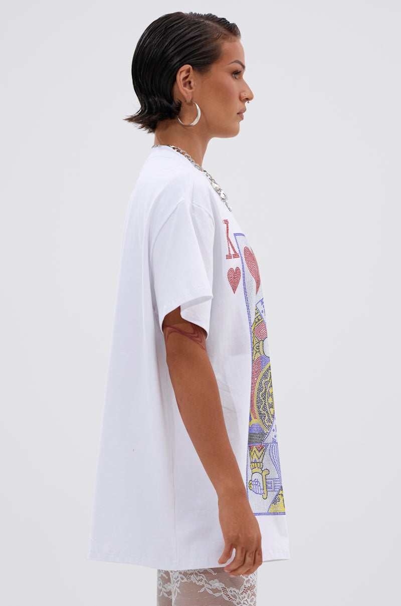 A woman with short, slicked-back hair and large hoop earrings stands in profile against a plain light background wearing the KING OF HEARTS OVERSIZED TEE IN WHITE, which features a colorful playing card design.