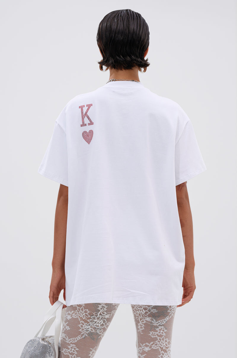 A person with short, dark hair is seen from behind wearing the KING OF HEARTS OVERSIZED TEE IN WHITE featuring a red "K" and heart graphic, sheer lace pants, and holding a silver textured bag.