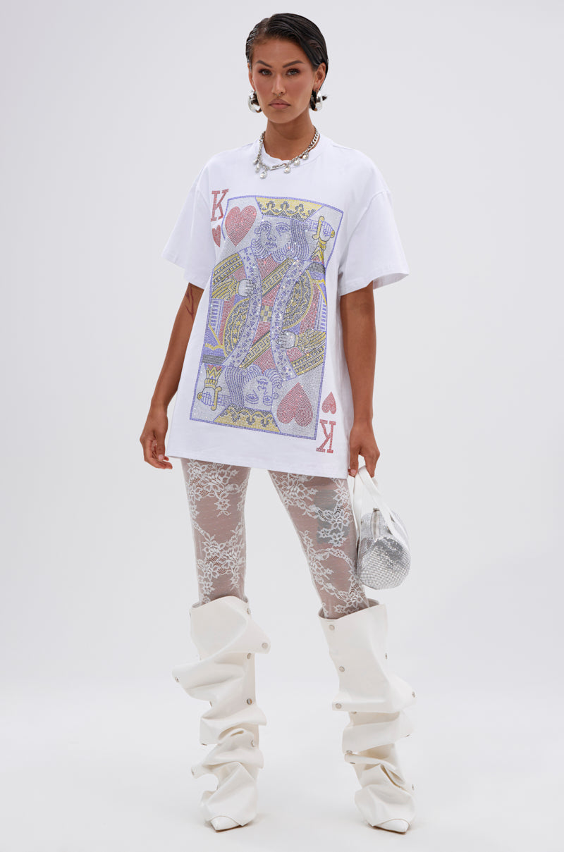 A woman stands confidently in the KING OF HEARTS OVERSIZED TEE IN WHITE by AKIRA Label, paired with sheer white lace leggings, tall slouchy white boots, silver earrings, and a small silver bag.