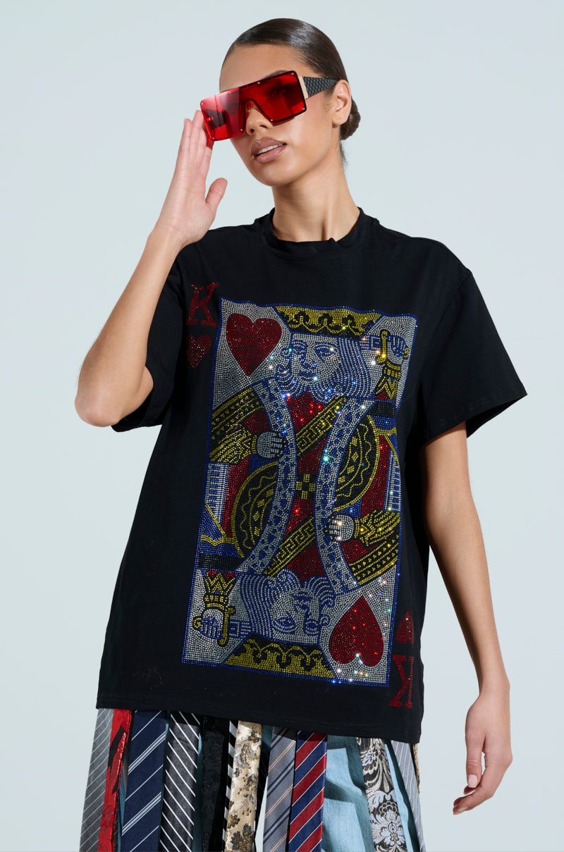 A woman in red sunglasses wears the KING OF HEARTS OVERSIZED TEE, featuring a rhinestone graphic of the King of Hearts and poker cards, paired with a skirt made from colorful neckties, against a plain background.