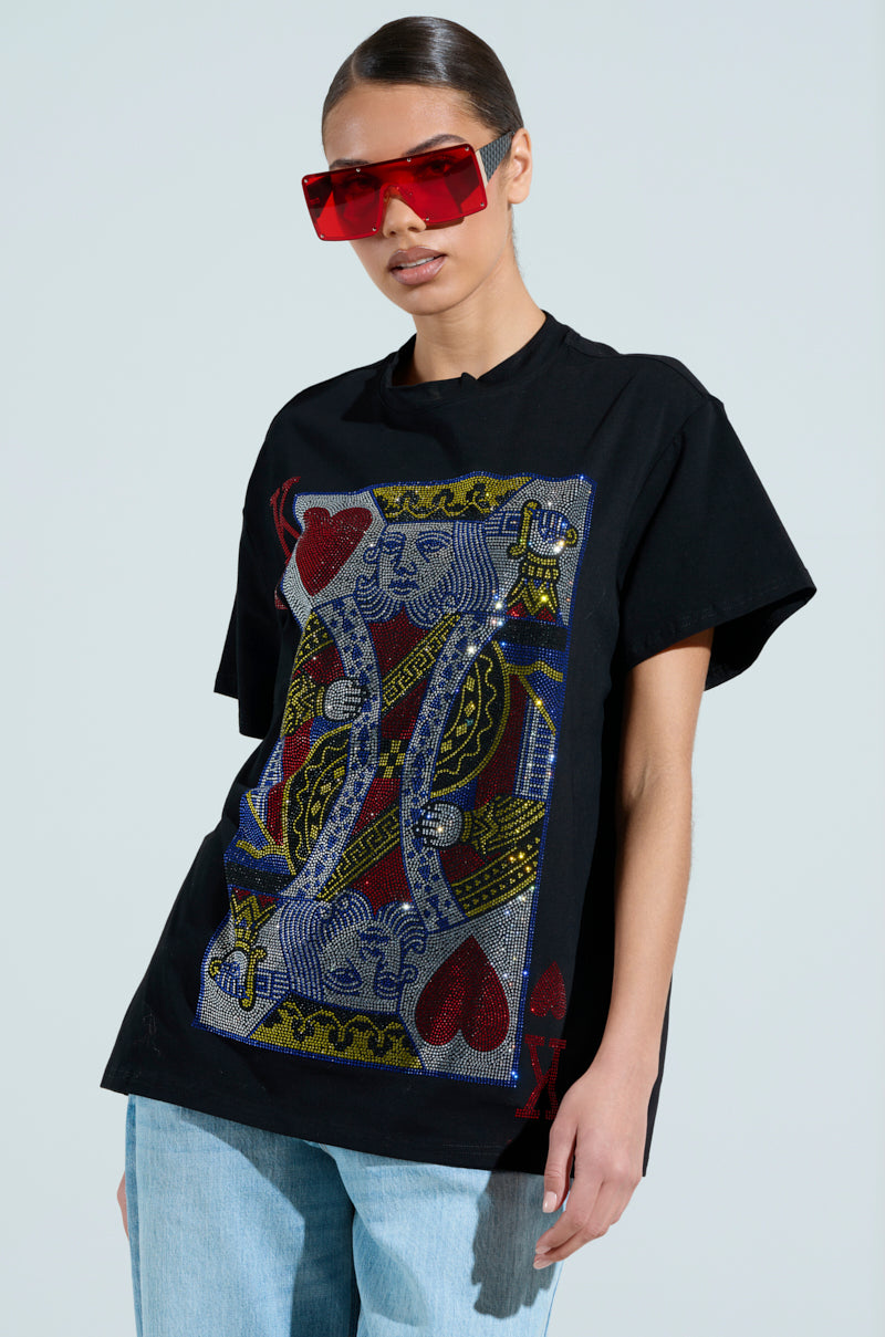 Wearing large red sunglasses and an oversized KING OF HEARTS OVERSIZED TEE featuring a rhinestone playing card graphic, they have short dark hair, light blue jeans, and are standing against a plain background.