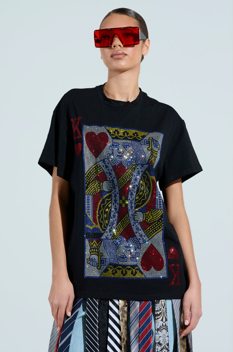 Wearing the KING OF HEARTS OVERSIZED TEE with a sequin design and large red sunglasses, they pair it with a skirt creatively made from neckties. The plain backdrop keeps all focus on this eclectic ensemble.