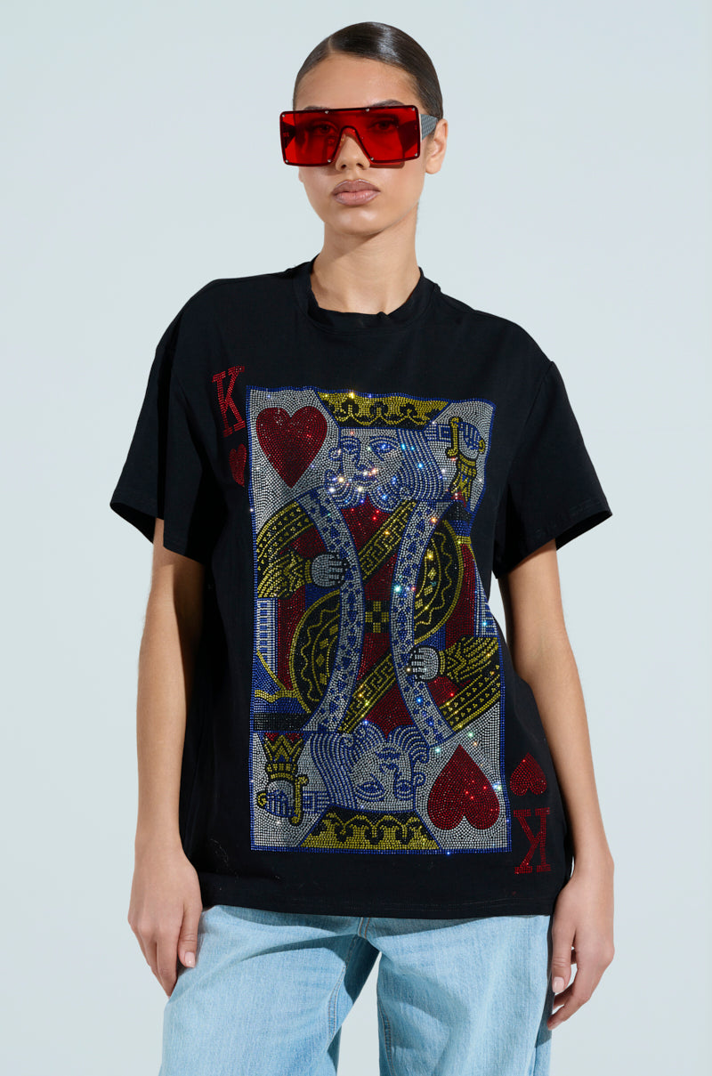 Donning oversized red sunglasses and the KING OF HEARTS OVERSIZED TEE, featuring a dazzling rhinestone King of Hearts design, someone casually poses with hands in pockets and light blue jeans against a plain backdrop.