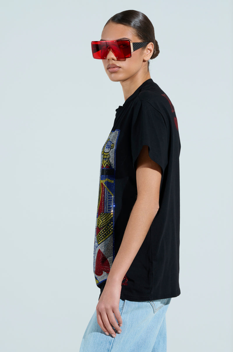 With hair tied back, a person wears oversized red sunglasses and the KING OF HEARTS OVERSIZED TEE, featuring a vibrant rhinestone graphic. Light blue jeans complete the ensemble against a plain light gray background.